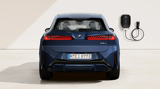 Auf dem Bild ist die Rückansicht eines weißen BMW iX3 zu sehen. Das Auto steht in einem Studio, und rechts davon ist eine schwarze BMW Wallbox, eine Ladestation, an der Wand befestigt. Die breiten, zweigeteilten Heckleuchten des Autos sind leuchtend rot und haben ein auffälliges, gestreiftes Design. In der Mitte der Heckklappe befindet sich das BMW-Emblem. Unterhalb des Nummernschilds ist eine dezente schwarze Kunststoffverkleidung. Durch die Heckscheibe sind die roten Sitze im Innenraum zu erkennen.