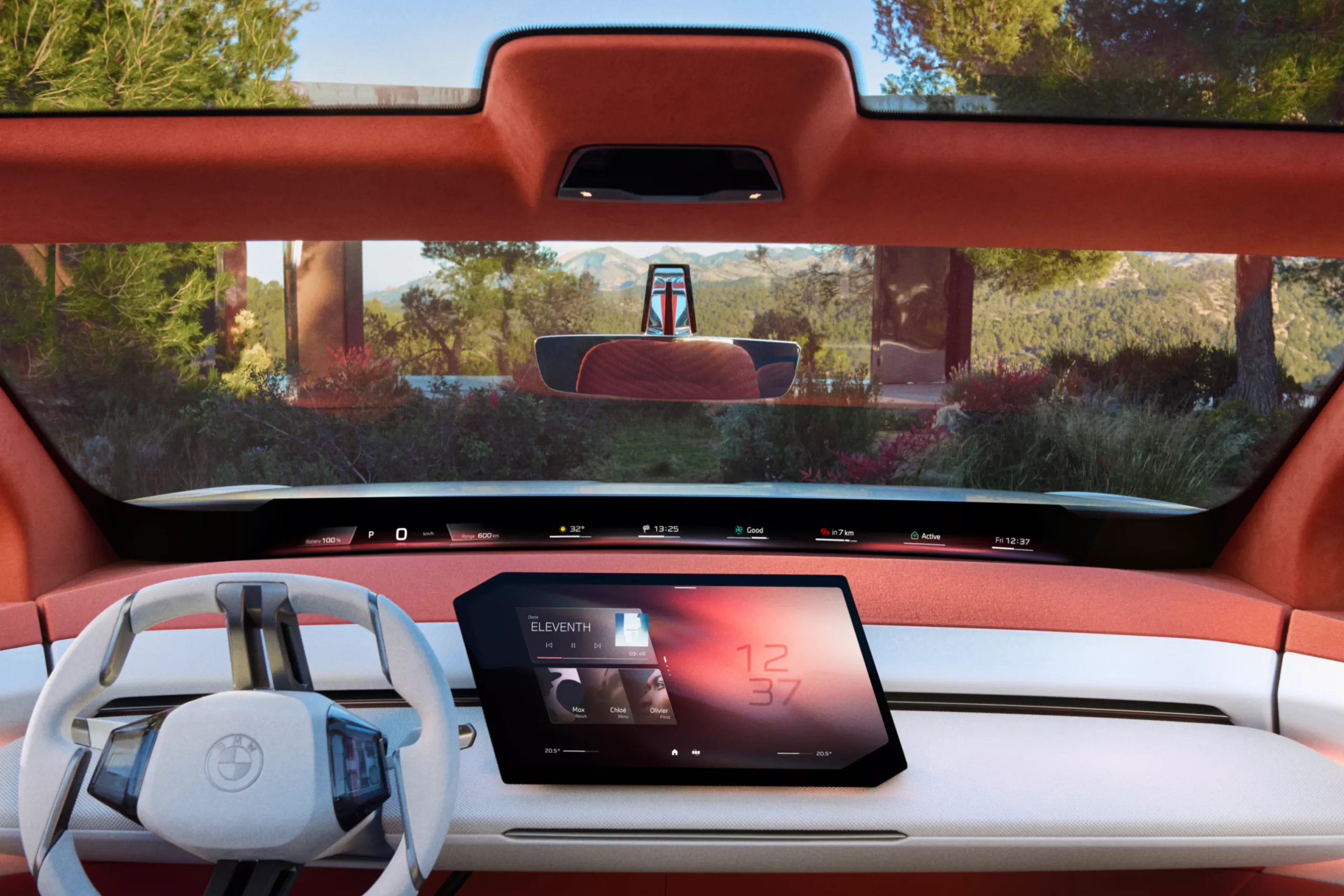 Das Bild zeigt den futuristischen Innenraum eines BMW-Konzeptautos. Das Cockpit ist minimalistisch und modern gestaltet, in Weiß- und Rottönen gehalten. Das Lenkrad ist weiß und hat ein zweispeichiges Design. Der obere Teil des Armaturenbretts und die Säulen sind mit einem orange-roten Stoff bezogen. Die untere Hälfte des Armaturenbretts ist weiß. Ein großes, rechteckiges Display in der Mitte dient als zentrales Bedienelement. Auf dem Bildschirm sind eine Uhrzeit, Musikinformationen und weitere Fahrzeugdaten zu sehen. Über die gesamte Breite des Armaturenbretts verläuft ein schmales Display, das Informationen wie Geschwindigkeit, Temperatur und Zeit anzeigt. Im Hintergrund, durch die Windschutzscheibe, ist eine sonnige Landschaft mit Bäumen und einem Gebäude zu erkennen.