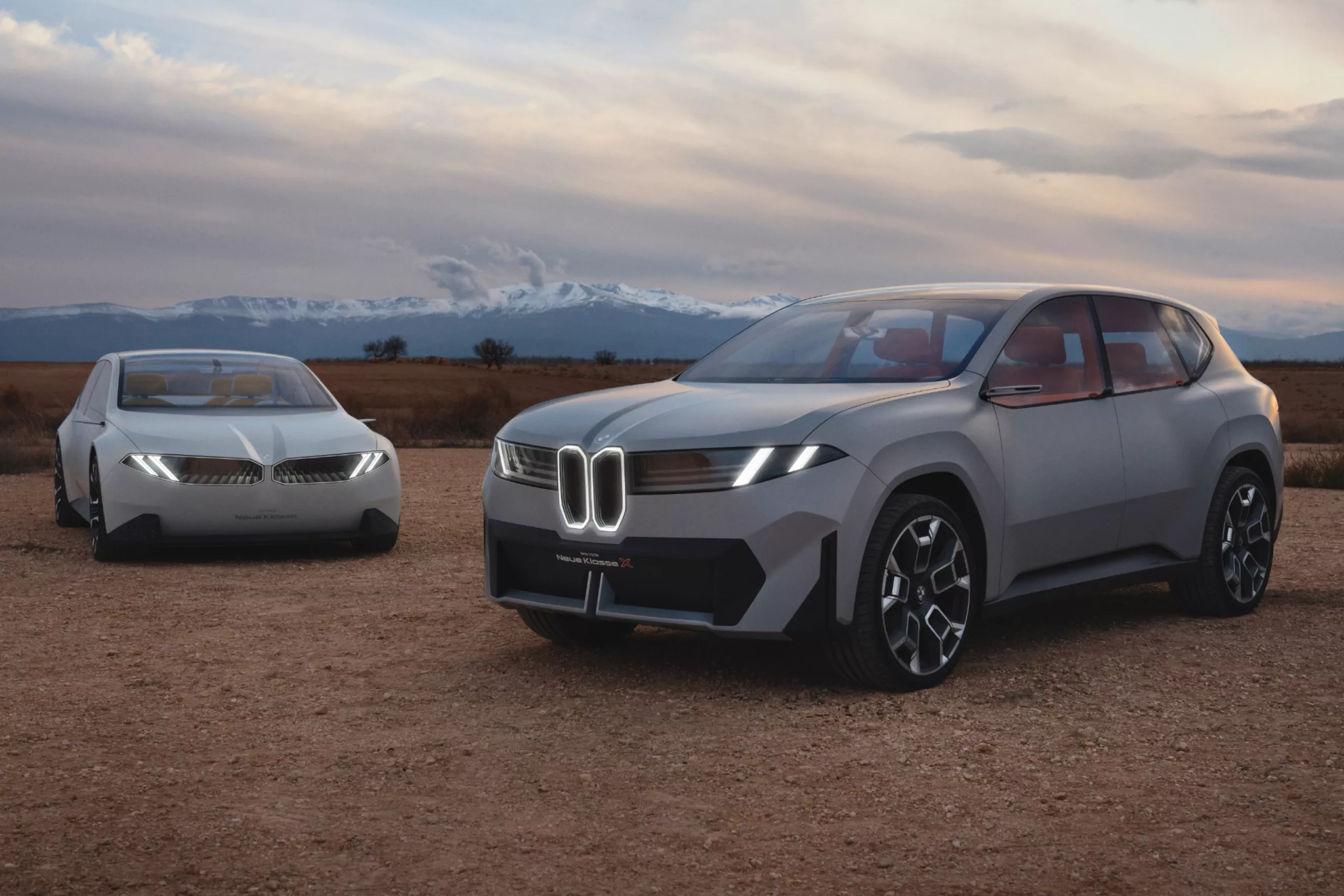Das Bild zeigt zwei moderne, weiße BMW-Konzeptautos in einer weiten, offenen Landschaft. Im Vordergrund ist ein SUV zu sehen, der einen großen Kühlergrill mit beleuchteten Elementen hat. Die Karosserie ist in einem hellen, matten Grau gehalten. Die Türen sind ohne sichtbare Griffe, und im Innenraum sind rote Sitze zu erkennen. Im Hintergrund steht ein weiteres, futuristisches Fahrzeug, das einer Limousine ähnelt. Der Untergrund ist ein unbefestigter, sandiger Boden. Im Hintergrund erstreckt sich eine weite Ebene bis zu einer schneebedeckten Bergkette. Der Himmel ist bewölkt, und die Beleuchtung deutet auf die Morgen- oder Abenddämmerung hin.