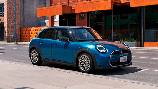 Ein blauer Mini mit schwarzem Dach und hellen Felgen ist von schräg vorne geparkt vor einem rötlichen Gebäude zu sehen.