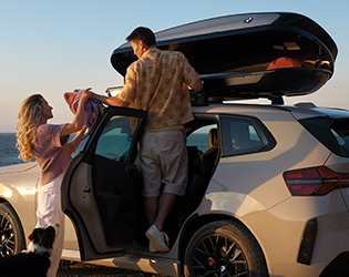 Ein Paar packt bei Sonnenuntergang am Meer eine geöffnete Dachbox auf einem beigen SUV. Eine Frau reicht dem auf dem Türschweller stehenden Mann farbige Decken nach oben. Ein Hund sitzt daneben.