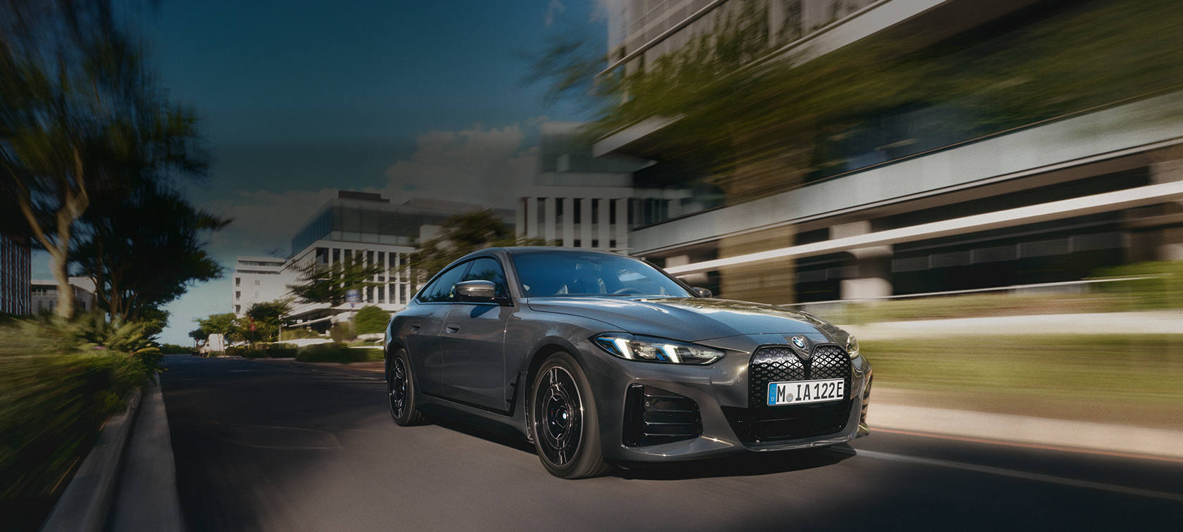 Eine dunkelgraue/anthrazitfarbene BMW-Limousine fährt dynamisch eine Straße entlang in einer modernen, städtischen Umgebung. Das Auto ist leicht nach vorne links geneigt und wird aus einer niedrigen Perspektive aufgenommen, was die Geschwindigkeit betont. Die Umgebung, bestehend aus modernen Bürogebäuden und Bäumen, ist durch die Bewegungsunschärfe der Aufnahme verschwommen, während das Auto scharf im Fokus steht. Das Licht deutet auf einen sonnigen Tag hin, möglicherweise am späten Nachmittag oder frühen Morgen.
