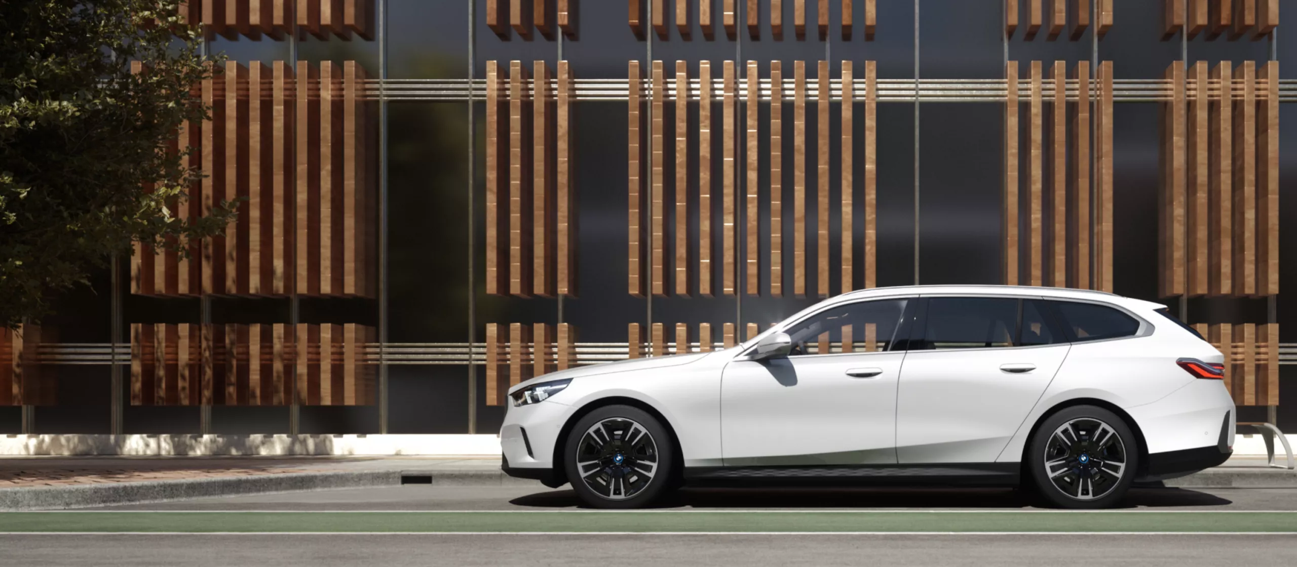 BMW 5er Touring Aktionsangebot Businesskunden Ein eleganter, weißer BMW Kombi parkt seitlich vor einem modernen Gebäude mit einer Glas- und Holzlamellenfassade. Links ist ein Teil eines Baumes zu sehen, rechts ein dunkles Dach