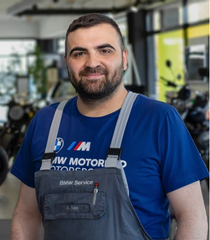 Ein Mann mittleren Alters mit dunklem Haar und Bart steht in einem Innenraum. Er trägt einen blauen BMW Motorrad Motorsport-Dienstleistungs-Overall über einem blauen T-Shirt und lächelt in die Kamera. Der Hintergrund ist unscharf, aber man kann ein Autohaus oder eine Werkstatt erkennen.