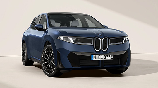 Auf dem Bild ist ein weißer BMW iX3 zu sehen, der frontal fotografiert wurde. Das Auto ist in einem Studio vor einem hellen Hintergrund platziert. Das Dach ist in kontrastierendem Schwarz gehalten. Die Frontpartie ist markant gestaltet, mit einer großen, rechteckigen "Doppel-Niere", die typisch für BMW ist, aber bei diesem Elektro-SUV geschlossen und schwarz ist. Die schlanken, horizontalen Scheinwerfer unterstreichen das moderne Design. Am unteren Teil der Frontstoßstange befinden sich schwarze Kunststoffelemente. Man kann auch durch die Scheiben die roten Sitze im Innenraum erkennen.