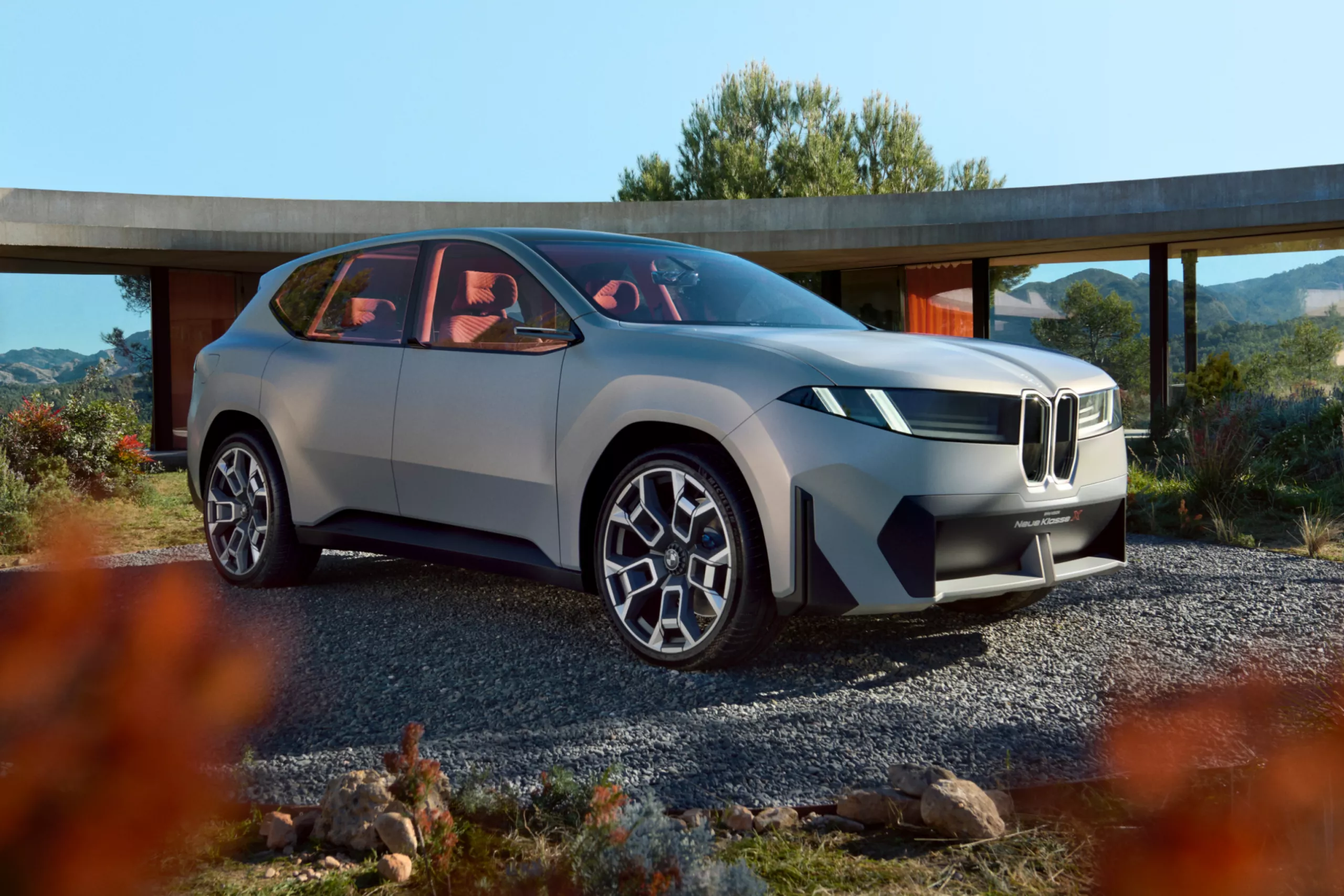 Das Bild zeigt ein graues, modernes BMW-Konzeptauto, das wie ein SUV aussieht. Es steht auf einem Kiesweg vor einem modernen Gebäude mit großen Glasflächen. Die Scheinwerfer sind horizontal und leuchten weiß. Der Kühlergrill ist vertikal und hat eine beleuchtete Umrandung. Im Inneren des Autos sind rote Sitze zu erkennen. Im Hintergrund sind Bäume und eine hügelige Landschaft zu sehen. Das gesamte Bild ist bei Tageslicht aufgenommen.
