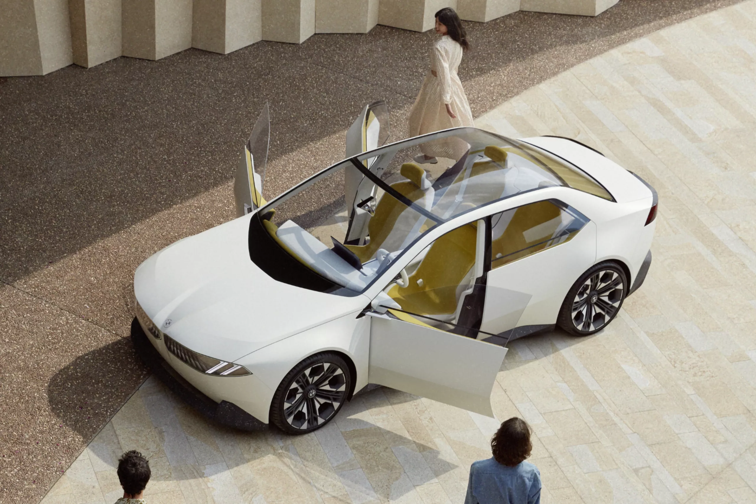 Das Bild zeigt einen weißen, modernen BMW, der von oben fotografiert wurde. Das Auto, eine Limousine, hat ein großes, durchsichtiges Dach und die Türen sind geöffnet. Der Innenraum des Fahrzeugs hat gelbliche Sitze. Eine Frau in einem hellen Kleid geht im Hintergrund am Auto vorbei, während im Vordergrund der Rücken einer weiteren Person zu sehen ist. Die Umgebung besteht aus hellen, geometrischen Fliesen und einem modernen Gebäude.