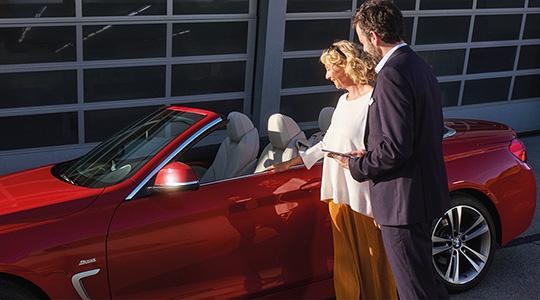 Ein BMW-Mitarbeiter im Anzug hilft einer lächelnden Kundin, die in gelber Hose und weißem Oberteil gekleidet ist, beim Einsteigen in ein rotes BMW 4er Cabrio mit offenem Verdeck vor einer Werkstatthalle