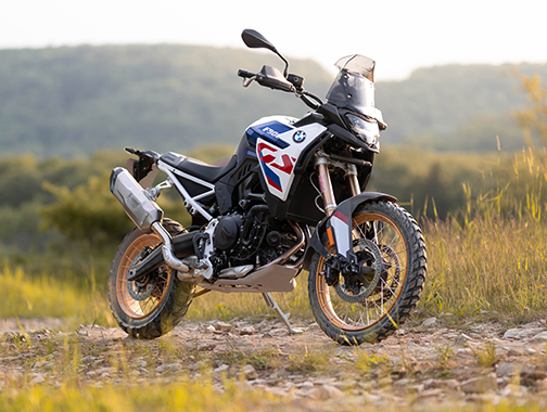  Auf dem Bild ist eine BMW F 900 GS abgebildet, ein großes, weiß-blau-rotes Motorrad, das auf einem unbefestigten Feldweg steht.  Das Motorrad ist in einem warmen, sonnigen Licht und steht seitlich zur Kamera auf einem Feldweg mit Grasbüscheln und Steinen. Im Hintergrund befinden sich bewaldete Hügel und eine bergige Landschaft. Es hat goldene Speichenräder, eine Verkleidung im BMW-Motorsportdesign in den Farben Weiß, Blau und Rot. Es ist sauber und glänzt, als wäre es brandneu.