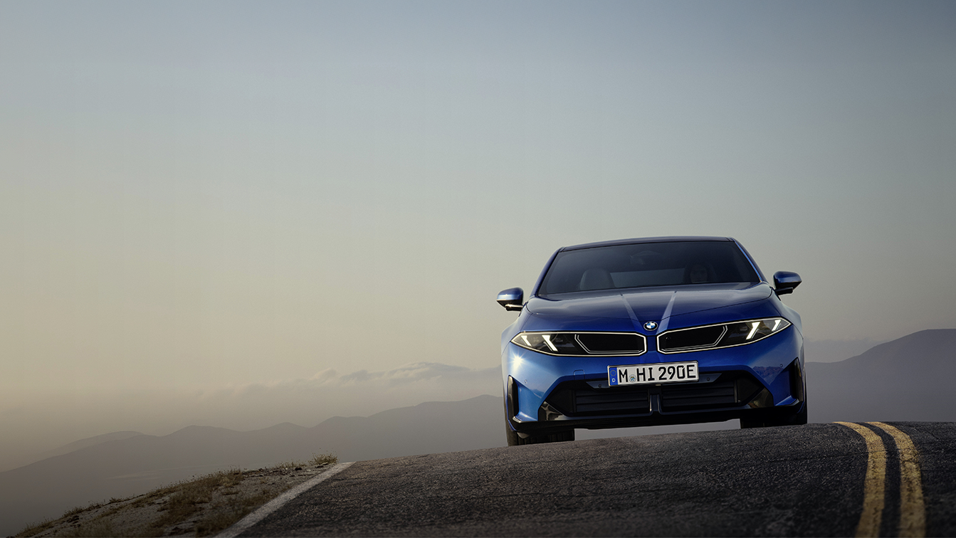 Neuer vollelektrischer BMW i3 in leuchtend blauer Lackierung aus der Frontperspektive auf einer Bergstraße. Die markante BMW Niere, die schmalen LED-Scheinwerfer und die sportlich gestaltete Frontpartie stehen im Mittelpunkt. Im Hintergrund erstreckt sich eine weitläufige Berglandschaft im sanften Licht der Abenddämmerung