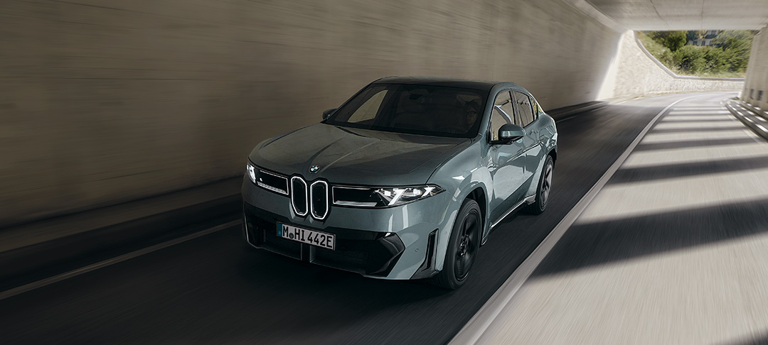 Neuer vollelektrischer BMW iX4 in graugrüner Lackierung während der Fahrt durch einen Tunnel mit markanter BMW Niere und LED-Scheinwerfern.