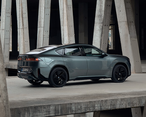 Graugrüner BMW iX4 in sportlicher Seiten- und Heckansicht mit schwarzen Leichtmetallrädern vor moderner Betonarchitektur.