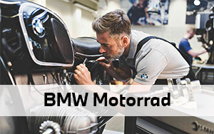 Das Bild zeigt einen Mechaniker in einer Werkstatt, der konzentriert an einem klassischen schwarzen Motorrad arbeitet. Er trägt graue Arbeitskleidung mit einem dezenten BMW-Logo am Ärmel und nutzt ein Werkzeug für feine Justierungen im Bereich unterhalb des Tanks.  Im Vordergrund ist der markante Boxermotor der Maschine zu erkennen, während im Hintergrund ein weiterer Mitarbeiter und Werkstattequipment angedeutet sind. Ein breiter, halbtransparenter weißer Balken liegt über der Mitte des Bildes und trägt den fetten schwarzen Schriftzug „BMW Motorrad“. Die gesamte Szene vermittelt einen Eindruck von handwerklicher Präzision und professionellem Service.