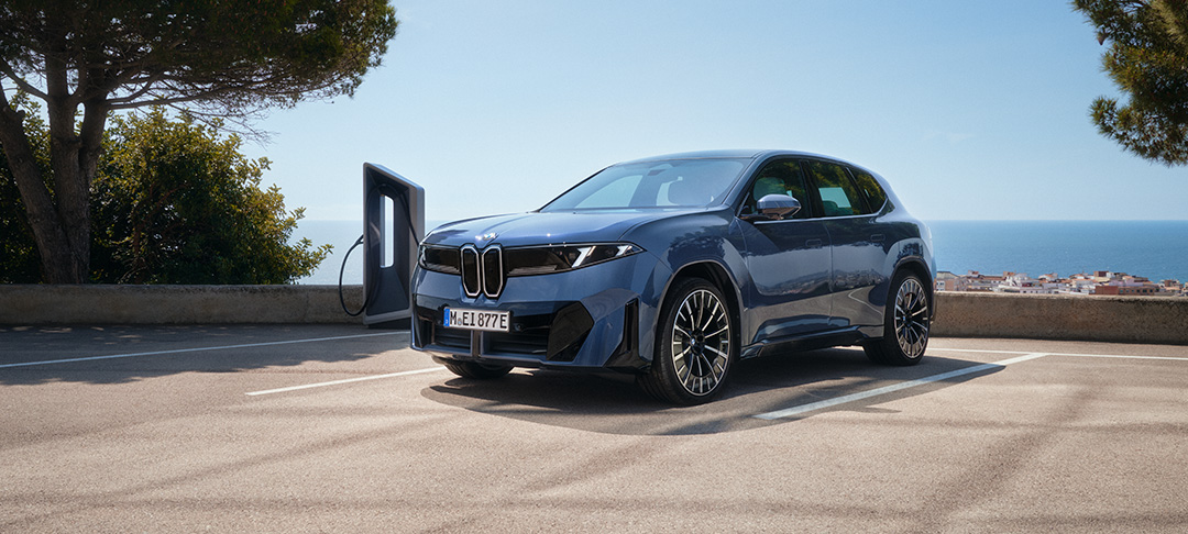 Blauer vollelektrischer BMW iX3 der Neuen Klasse an einer Ladestation mit Blick auf das Meer und eine Küstenlandschaft