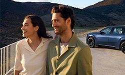 Lächelndes Paar vor einer Berglandschaft mit einem blauen BMW der Neuen Klasse im Hintergrund