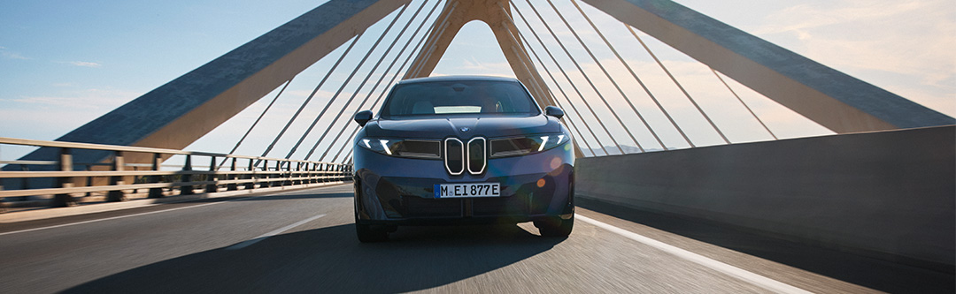 Blauer BMW iX3 der Neuen Klasse in dynamischer Frontansicht während der Fahrt über eine moderne Schrägseilbrücke
