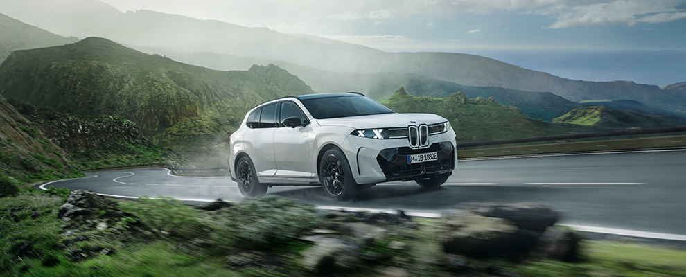 Weißer BMW X5 50e xDrive während der Fahrt auf einer kurvenreichen Bergstraße mit grüner Landschaft und markanter BMW Niere.