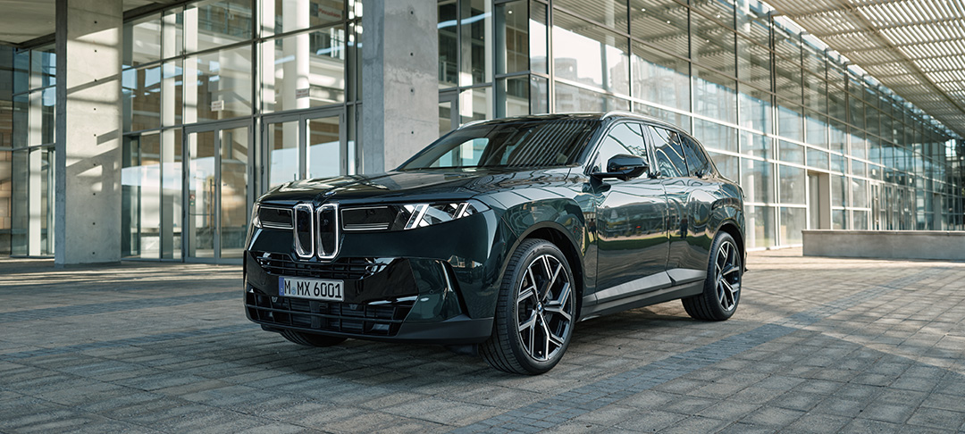 Neuer BMW X5 in dunkler Lackierung aus der vorderen Seitenperspektive vor einem modernen Gebäude mit großer Glasfassade. Die markante BMW Niere, die schmalen LED-Scheinwerfer und die großen Leichtmetallräder prägen das kraftvolle SUV-Design.