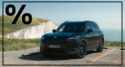 Ein schwarzer, vollelektrischer SUV, der auf einem Schotterparkplatz an der Küste steht. Im Hintergrund sieht man eine markante weiße Steilküste, grünes Gras und das weite, blaue Meer unter einem klaren Himmel. Das Auto wirkt abenteuerlustig und großzügig. Auch hier ist oben links ein markantes, schwarzes Prozentzeichen eingeblendet.
