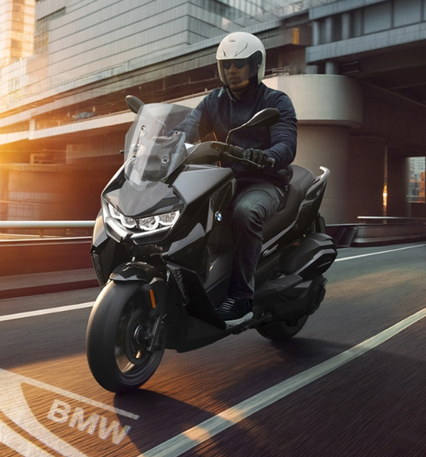 Ein Mann in dunkler Kleidung und einem weißen Integralhelm fährt auf einem modernen, mattschwarzen BMW Maxi-Scooter, erkennbar an der prominenten Frontverkleidung und der großen Windschutzscheibe. Der Roller bewegt sich auf einer städtischen Straße mit Hochhäusern im Hintergrund. Das Licht der tiefstehenden Sonne scheint intensiv von links ins Bild und wirft warme Schatten. Im Vordergrund ist der Schriftzug "BMW" als helle Markierung auf den dunklen Asphalt gemalt.