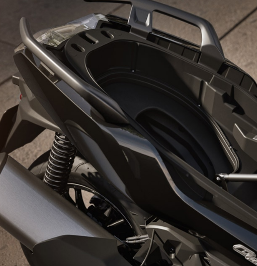 Detailaufnahme der Heckpartie eines glänzend schwarzen Motorrollers mit sichtbarem Stauraum unter dem hochgeklappten Sitz. Der Stauraum ist eine tiefe, schwarze Mulde mit ovalen Ausschnitten. Links im Bild dominiert der silberne, mattierte Auspuff mit einem breiten Endrohr. Darüber sind die silberne Haltestange für den Beifahrer sowie die offenliegende, spiralförmige Federung des Hinterrads gut erkennbar. Der Fokus liegt auf der Funktionalität und dem robusten Design des hinteren Fahrzeugteils.