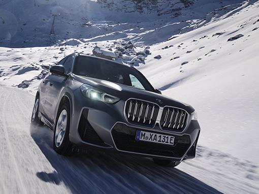 Ein grauer BMW X1 fährt auf einer schneebedeckten Straße bergauf, die Sonne blendet leicht von rechts