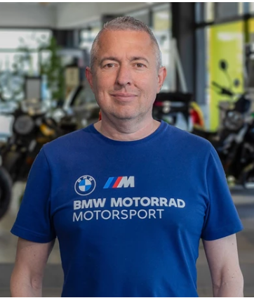 Das Bild zeigt einen Mann mittleren Alters mit kurzen, grau melierten Haaren, der direkt in die Kamera blickt. Er trägt ein dunkelblaues T-Shirt, auf dem in weißen Buchstaben „BMW MOTORRAD MOTORSPORT“ und das BMW-Logo zu sehen sind. Der Hintergrund ist unscharf und zeigt eine Motorrad-Ausstellungshalle.