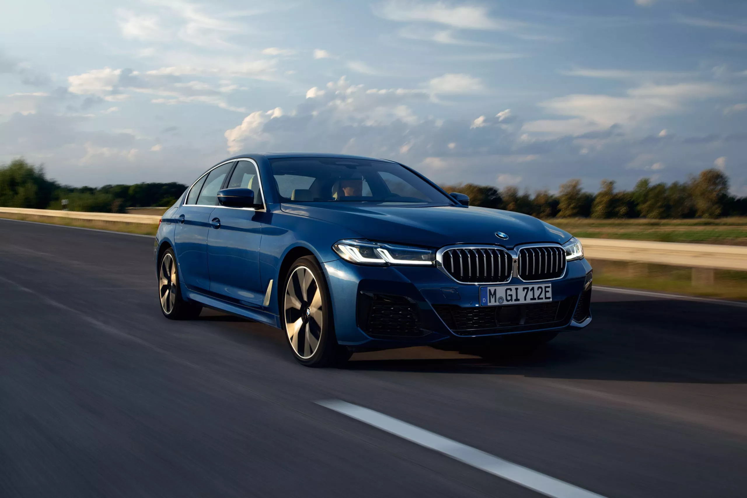  Dunkelblauer BMW fährt dynamisch auf einer Straße mit bewölktem Himmel.