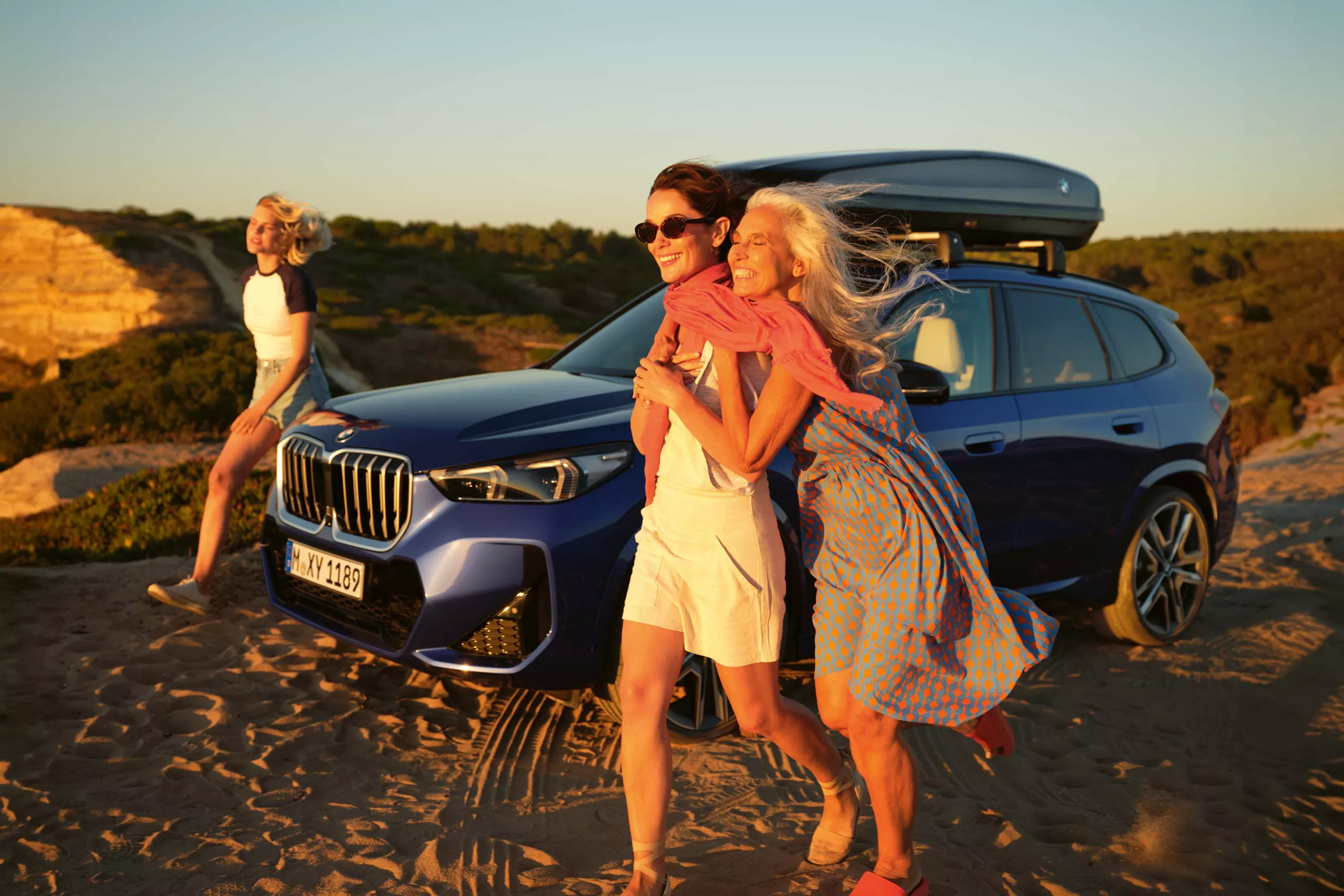 Drei Frauen am Strand neben einem blauen BMW SUV mit Dachgepäckträger, der auf Sand parkt.