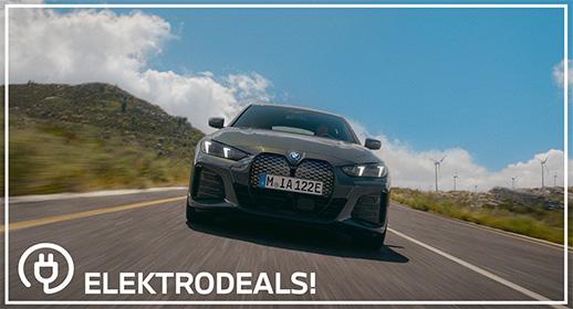 Vorderansicht einer grauen BMW i4 Elektro-Limousine mit Kennzeichen 'MIA 122E', die auf einer Bergstraße an Windrädern vorbeifährt. Mit 'Elektrodeals!'-Logo."