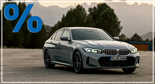 Hellgraue BMW 318i Limousine parkend auf einem Asphaltplatz vor Gebirgssilhouette in der Dämmerung; blaues Prozent-Symbol oben links.