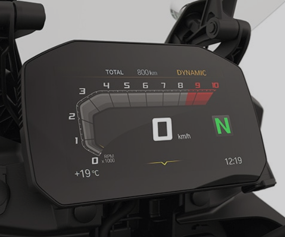 Nahaufnahme eines modernen, digitalen Motorrad-Cockpits mit schwarzem Hintergrund. Das Display zeigt eine digitale Tachonadel für die Drehzahl (RPM) mit einer Skala von 0 bis 10 und einem roten Bereich bei 9 und 10. In der Mitte steht die aktuelle Geschwindigkeit von 0 km/h in großen, weißen Ziffern. Rechts daneben leuchtet ein großes, grünes "N" für den Leerlauf. Am oberen Rand sind der Gesamtkilometerstand von 800 km und der aktive Fahrmodus "DYNAMIC" in gelb zu sehen. Weitere Anzeigen umfassen die Außentemperatur von +19 °C unten links und die Uhrzeit 12:19 unten rechts.