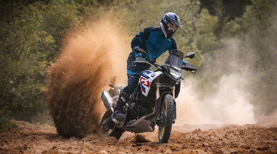 Ein Motorradfahrer in blauer und schwarzer Offroad-Kleidung und passendem Helm fährt ein weiß-blau-rotes BMW Adventure-Motorrad dynamisch durch einen schlammigen Untergrund. Die Aufnahme fängt den Moment ein, in dem das Hinterrad des Motorrads große Mengen an braunem Schlamm und Erde in einer hohen Fontäne aufwirbelt. Der Fahrer neigt sich nach innen, während das Motorrad schräg durch die Kurve fährt. Im Hintergrund ist ein dichter, grüner Wald oder Bewuchs zu sehen. Das Bild vermittelt Geschwindigkeit und die Eignung des Motorrads für unwegsames Gelände.