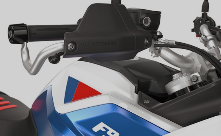 Nahaufnahme der Lenkerpartie eines Motorrads, die den Handschutz, den Lenker und einen Teil des weiß-blau-roten Tanks zeigt. Ein massiver, schwarzer Handschutz mit dem Schriftzug "BMW Motorrad" in der Mitte ist an dem silbernen Lenker befestigt. Der vordere Teil des Tanks ist in den Farben Weiß und Blau gehalten, mit einem roten Dreieck als Akzent. Der schwarze Griff des Lenkers ist sichtbar, ebenso wie die Bremsflüssigkeitsbehälter und verschiedene Kabel. Die Gesamtästhetik ist sportlich und technisch.