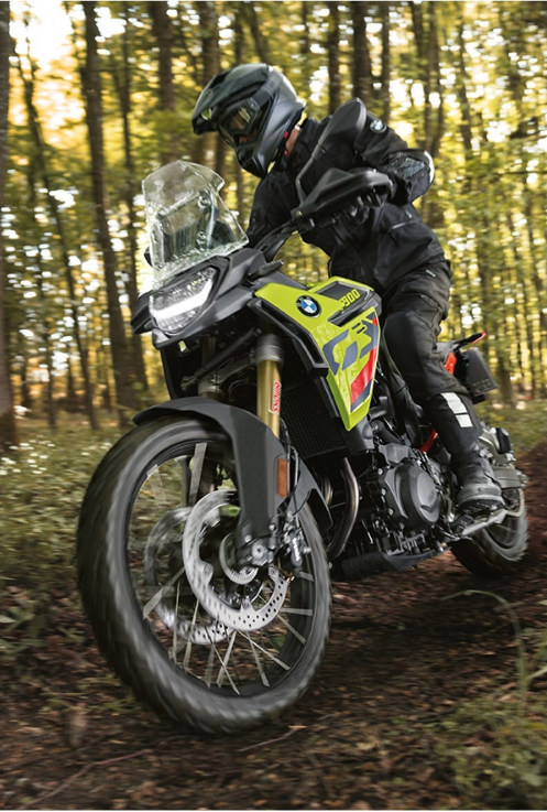 Ein Motorradfahrer in schwarzer Offroad-Ausrüstung und dunklem Helm fährt dynamisch auf einem dunkelbraunen Waldweg, umgeben von dichtem Baumbestand. Er sitzt auf einem geländegängigen BMW Adventure-Motorrad, das eine auffällige, leuchtend neongelbe Seitenverkleidung mit schwarzen und roten Grafiken besitzt. Das Motorrad hat eine hohe Windschutzscheibe, robuste Handschützer und eine sichtbare goldene Federung. Die Aufnahme ist aus niedriger Perspektive gemacht, wobei das Vorderrad und die Speichenbremse im Vordergrund dominieren und die Geschwindigkeit der Fahrt im schattigen Wald betonen.