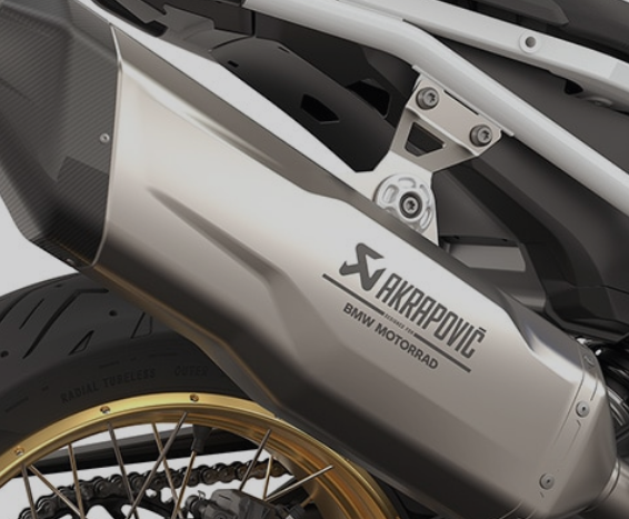 Nahaufnahme eines großen, silbernen Motorradauspuffs der Marke Akrapovič, der schräg von unten nach oben verläuft. Der Auspuff ist zylindrisch, aus mattem Metall und trägt den Schriftzug "AKRAPOVIČ" zusammen mit dem BMW Motorrad Logo auf seiner Seite. Das Ende des Auspuffs ist mit einer dunklen, Kohlefaser-Endkappe versehen. Im unteren linken Bereich ist ein Teil des goldfarbenen Speichenrads des Motorrads und die Antriebskette zu erkennen. Die gesamte Baugruppe ist an einem weißen Rahmen befestigt, der im oberen Teil des Bildes sichtbar ist.