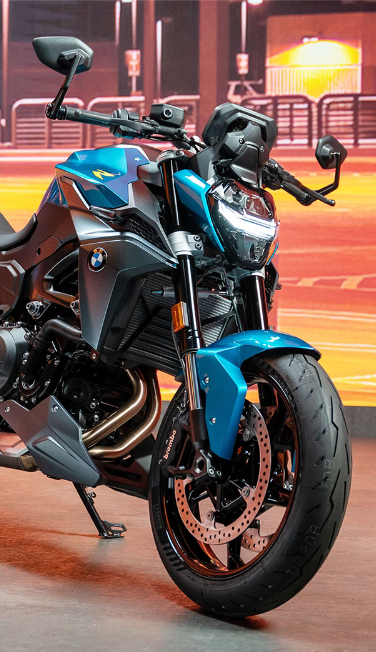 Nahaufnahme der Front eines sportlichen BMW Motorrads, das vor einem unscharfen, beleuchteten städtischen Hintergrund steht. Das Motorrad ist in einem auffälligen Türkisblau lackiert, das mit matten Grau- und Schwarztönen an Motor und Verkleidung kontrastiert. Die Frontansicht wird von dem modernen LED-Scheinwerfer und dem breiten Vorderrad dominiert, dessen gelochte Scheibenbremsen und die Bremssättel der Marke Brembo gut sichtbar sind. Der glänzende Krümmer des Motors verläuft im unteren linken Bereich des Bildes. Die gesamte Szene ist durch das orange-gelbe Licht des Hintergrunds beleuchtet.