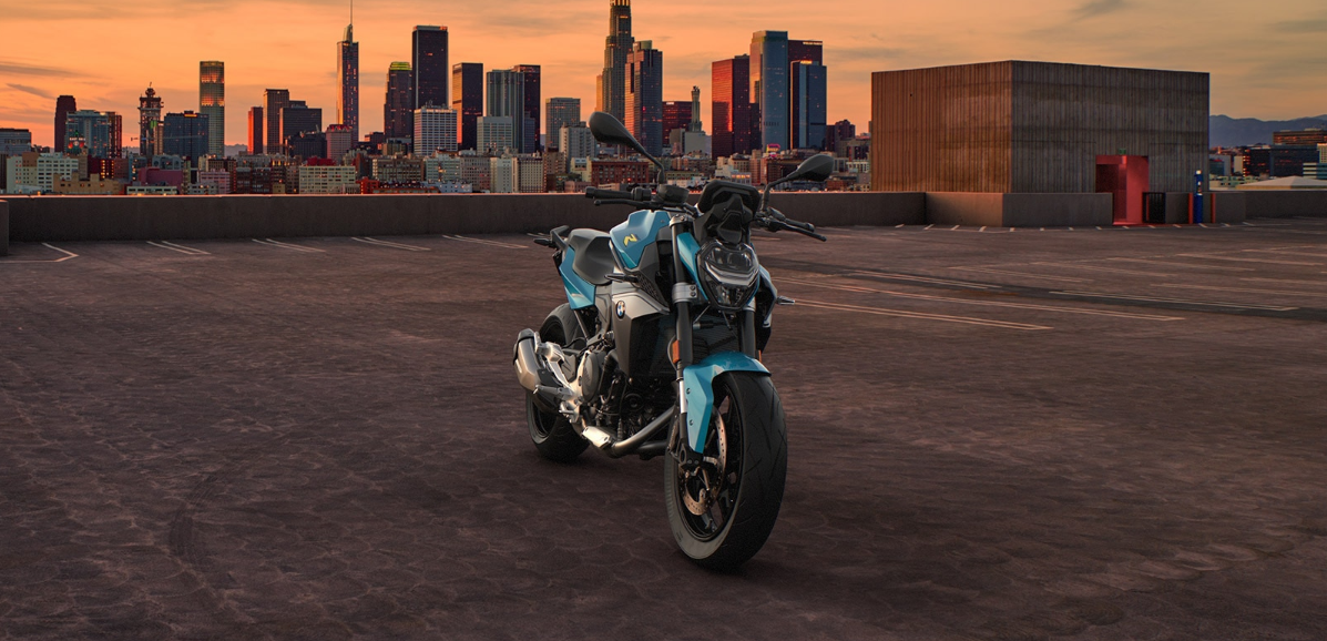 Ein modernes, blau-weißes BMW Naked Bike (Motorrad ohne Verkleidung) steht zentral auf einem großen, leeren Parkdeck im Sonnenuntergang. Die Front des Motorrads ist direkt auf den Betrachter gerichtet. Im Hintergrund erstreckt sich die Skyline einer Großstadt mit hohen Hochhäusern, deren Fenster von der orange-roten Abendsonne beleuchtet werden. Rechts begrenzt ein massives, kastenförmiges Gebäude das Parkdeck. Das Motorrad, das Parkdeck und die Gebäude sind in das warme Licht der Dämmerung getaucht.