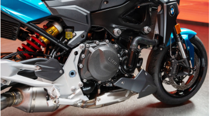 Nahaufnahme des Motors und der Aufhängung eines sportlichen BMW Motorrads, das von der Seite gezeigt wird. Das Zentrum dominiert die dunkelgraue Motorabdeckung mit dem BMW-Schriftzug. Im unteren Bereich verläuft eine silberne Auspuffanlage mit einem hellen Endschalldämpfer. Darüber ist die rot gewickelte Feder des Federbeins sichtbar, dessen Gehäuse goldgelb abgesetzt ist. Der obere Bereich zeigt die türkisblaue Seitenverkleidung und den schwarzen Motorrahmen. Der Vorderreifen ist am rechten Bildrand zu sehen.