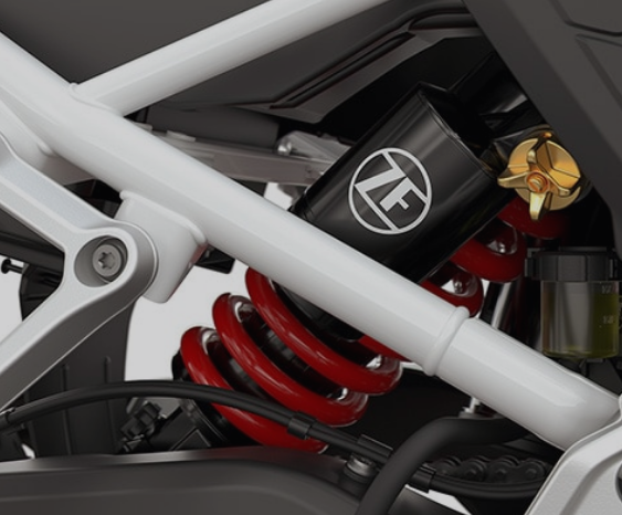 Nahaufnahme der Hinterradaufhängung eines Motorrads, die von einem weißen Rahmen umgeben ist. Im Zentrum liegt ein Stoßdämpfer mit einem schwarzen Gehäuse, auf dem das weiße ZF-Logo abgebildet ist. Die sichtbare Feder ist leuchtend rot und wickelt sich um den Dämpfer. Rechts oben ist das goldfarbene Einstellrad des Dämpfers sowie ein kleiner Behälter mit gelber Flüssigkeit (Bremsflüssigkeit) zu erkennen. Die gesamten mechanischen Komponenten sind sauber und kontrastieren scharf mit dem hellen Hintergrund.