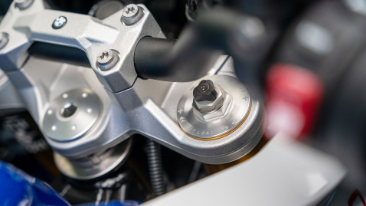 Nahaufnahme der oberen Gabelbrücke und Lenkerbefestigung eines Motorrads, die alle aus hellem, mattem Aluminium gefertigt sind. Der Lenker ist ein schwarzes Rohr, das in silbernen Klemmen gehalten wird. Im rechten Vordergrund ist das obere Ende des Federbeins mit einer schwarzen Einstellschraube zu sehen. Die Oberfläche des Aluminiumteils ist fein bearbeitet, und im unteren rechten Bereich ist die weiße Verkleidung des Motorrads sichtbar, die scharf mit dem technischen Element kontrastiert.