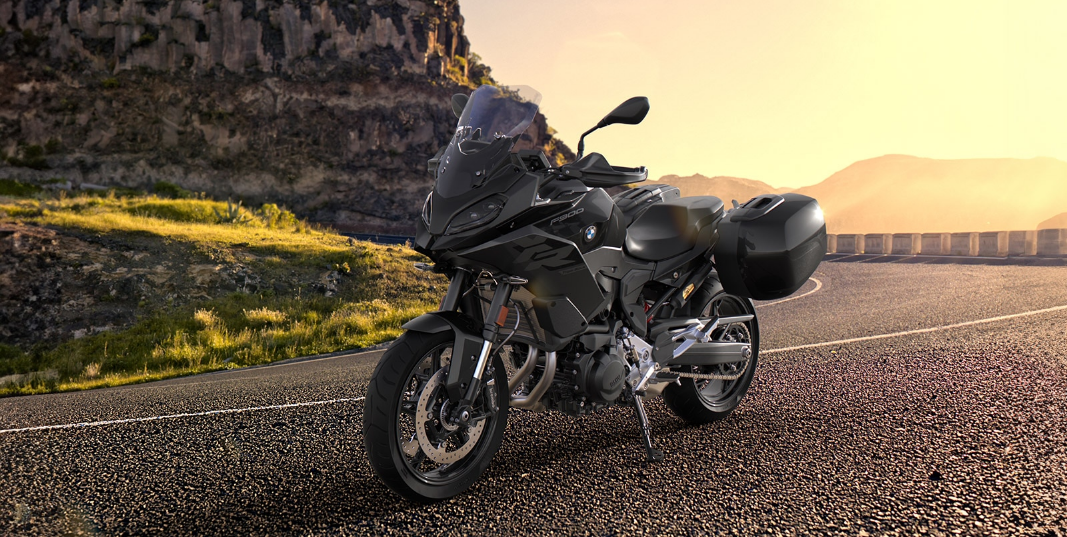 Ein tiefschwarzes BMW Adventure-Motorrad steht auf dem Asphalt einer kurvigen Bergstraße, die von der untergehenden Sonne beleuchtet wird. Das Motorrad ist mit einer hohen Frontscheibe und zwei schwarzen Seitenkoffern für Reisen ausgestattet. Es steht auf seinem Seitenständer und ist von vorne leicht rechts aufgenommen. Im Hintergrund erstreckt sich eine felsige, braune Berglandschaft. Das warme, goldene Licht des Sonnenuntergangs scheint von rechts über die Straße und die Berge.