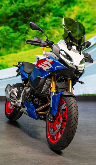 Ein BMW Adventure-Motorrad, das in den auffälligen Farben Blau, Weiß und Rot lackiert ist, steht auf einem dunklen Boden vor einem unscharfen, grünen Hintergrund. Die Front des Motorrads wird von einer weißen Verkleidung und einer hohen, klaren Windschutzscheibe dominiert. Besonders hervorstechend sind die roten Felgen und die gelochten Bremsscheiben an den Vorderrädern. Das Design wirkt sportlich und reisebereit, mit goldfarbenen Federbeinen an der Front. Die blauen und roten Grafiken an den Seitenverkleidungen betonen den dynamischen Look des Motorrads.