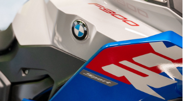 Nahaufnahme der Tank- und Seitenverkleidung eines sportlichen BMW Motorrads. Die Verkleidung ist oben glänzend weiß und trägt das runde BMW-Emblem in der Mitte. Am oberen Rand ist in roter Schrift "F 900" zu erkennen. Im unteren Bereich schließt eine blau lackierte Seitenverkleidung an, die einen großen weißen Grafikstreifen und den kleinen Schriftzug "Sport" trägt. Das gesamte Detail wirkt dynamisch und mehrfarbig.
