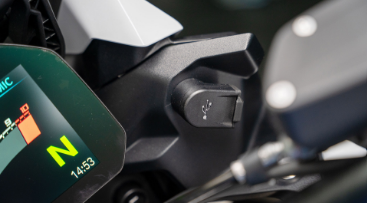 Nahaufnahme eines technischen Details am Cockpit eines Motorrads, das eine Ladebuchse zeigt. Zentral im Bild ist ein schwarzes Kunststoffgehäuse mit einer USB-Ladebuchse zu sehen. Links daneben ist ein Teil des digitalen Displays sichtbar, das den Leerlauf "N" und die Uhrzeit "14:53" anzeigt. Darüber befindet sich ein Teil der weißen Verkleidung. Die gesamte Szene ist ein Detail, das die Konnektivitäts-Funktionen des Motorrads hervorhebt.