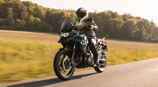 Ein Motorradfahrer in olivgrüner Motorradkleidung und geschlossenem, schwarz-weißem Helm fährt auf einem dunkelblauen BMW-Adventure-Motorrad, der GS-Serie, auf einer schmalen Landstraße. Das Motorrad hat eine hohe Frontscheibe und fährt dynamisch von vorne auf den Betrachter zu. Der Hintergrund zeigt links und rechts goldgelbe Felder, die sich bis zu einem dichten, grünen Waldrand erstrecken. Die Szene ist in warmes Licht getaucht, was eine friedliche, schnelle Fahrt bei Sonnenschein vermittelt.