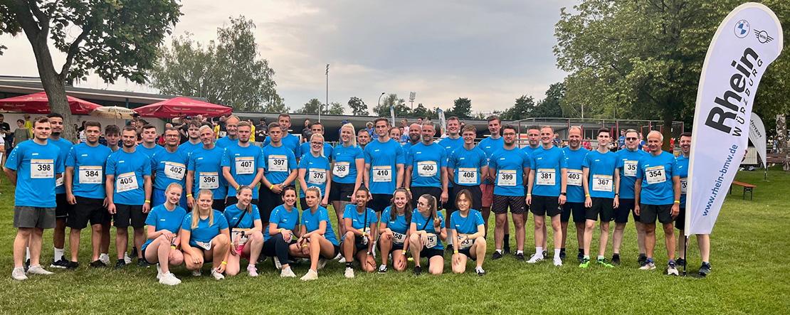 BMW Rhein Firmenlauf Würzburg Große Gruppe von Personen in blauen T-Shirts mit Startnummern posiert auf einer Wiese mit Rhein Gruppe Flagge