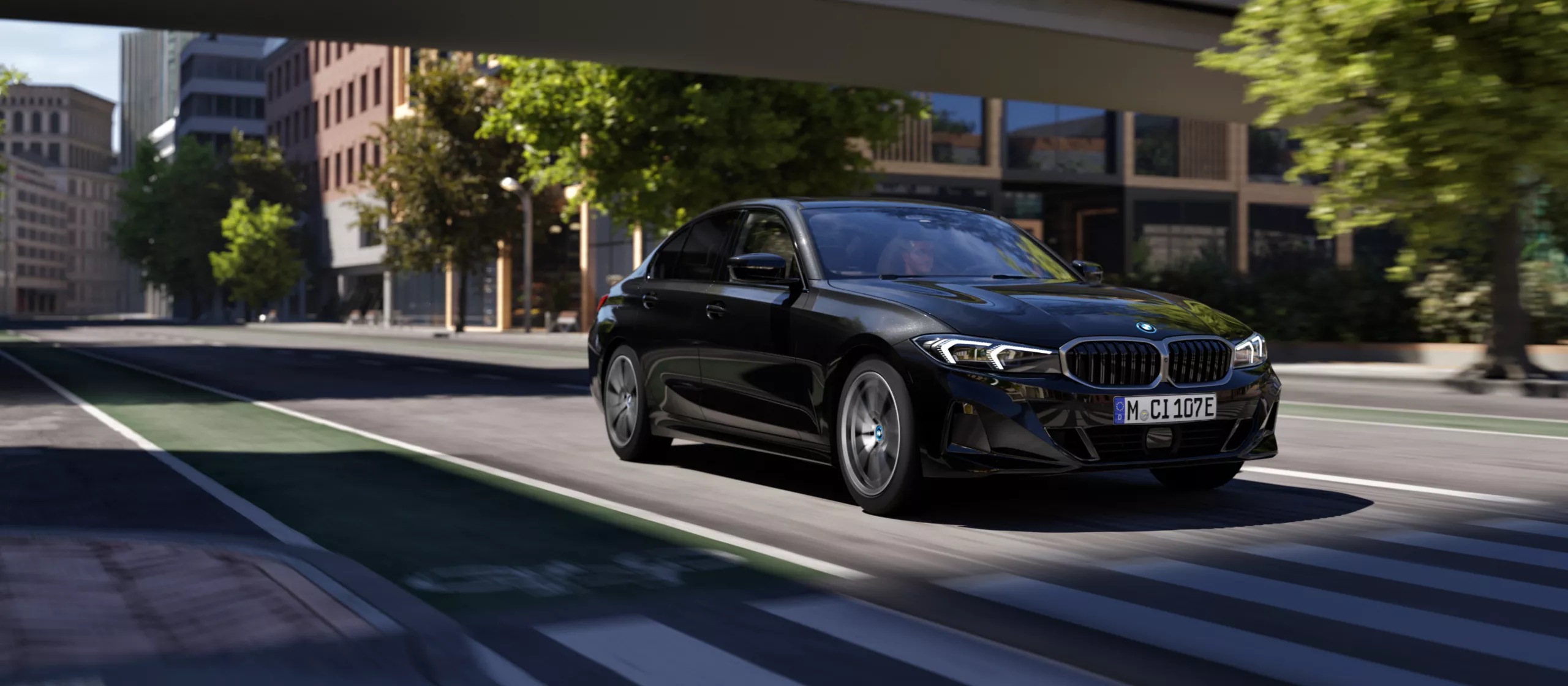 Das Bild zeigt eine schwarze BMW Limousine (vermutlich ein Modell der 3er-Reihe) in dynamischer Fahrt.  Das Auto fährt auf einer breiten Stadtstraße mit einer grün markierten Spur (wahrscheinlich ein Radweg oder eine Busspur) und überquert gerade einen Zebrastreifen. Die Aufnahme ist aus einer niedrigen Perspektive und leicht von vorne gemacht, was dem Bild viel Geschwindigkeit und Bewegung verleiht.  Im Hintergrund sind eine moderne Stadtkulisse mit mehrstöckigen Gebäuden aus Ziegeln und Glas sowie Bäumen entlang der Straße zu sehen. Die Beleuchtung ist hell und suggeriert Tageslicht. Das Kennzeichen des Wagens ist mit "M CI 107E" lesbar.