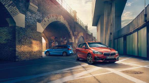BMW Gebrauchtwagen Service Inclusive Ein roter BMW 2er Active Tourer steht auf einem sonnigen Platz unter einer gemauerten Brücke mit Bögen. Im Hintergrund, weiter hinten unter einem anderen Bogen, ist ein blauer BMW zu sehen. Die Szene hat eine städtische, leicht industrielle Atmosphäre mit starkem Gegenlicht.