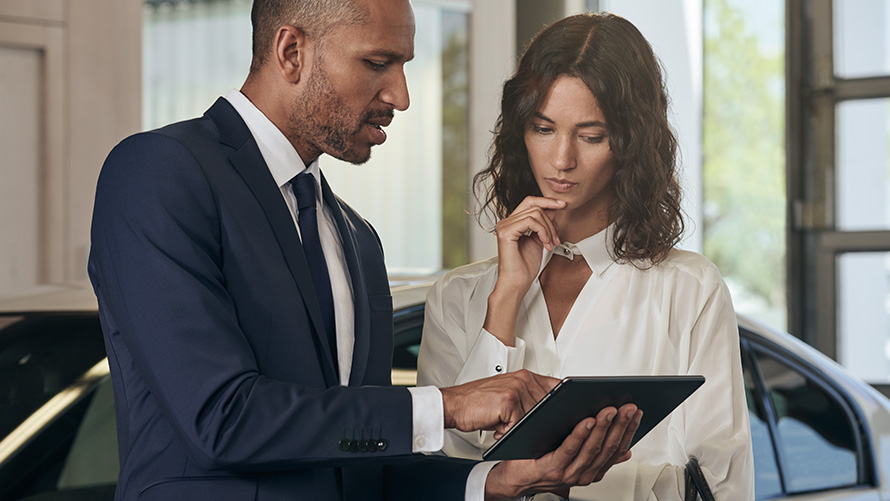 Das Motiv zeigt ein professionelles Beratungsgespräch zwischen einem BMW-Mitarbeiter und einer Kundin direkt am Fahrzeug. Beide betrachten konzentriert ein Tablet, was die moderne, digitale Konfiguration von Dienstwagen und die individuelle Betreuung von Geschäftskunden bei der Rhein Gruppe symbolisiert.